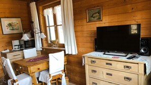 A wooden cabin interior with a wooden dining table, white chairs, a TV, a wooden chest of drawers, and a window with curtains. - Rotes Haus Füssen Apartment Romantik (Füssen)