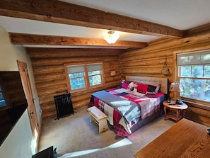 Quarto de cabana de toras aconchegante, com uma cama grande, um banquinho, uma mesa pequena e uma janela.