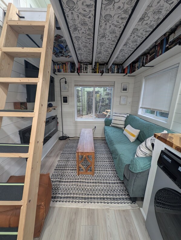 A compact living space with a loft bed, a small table, a couch, a window, and a bookshelf filled with books.