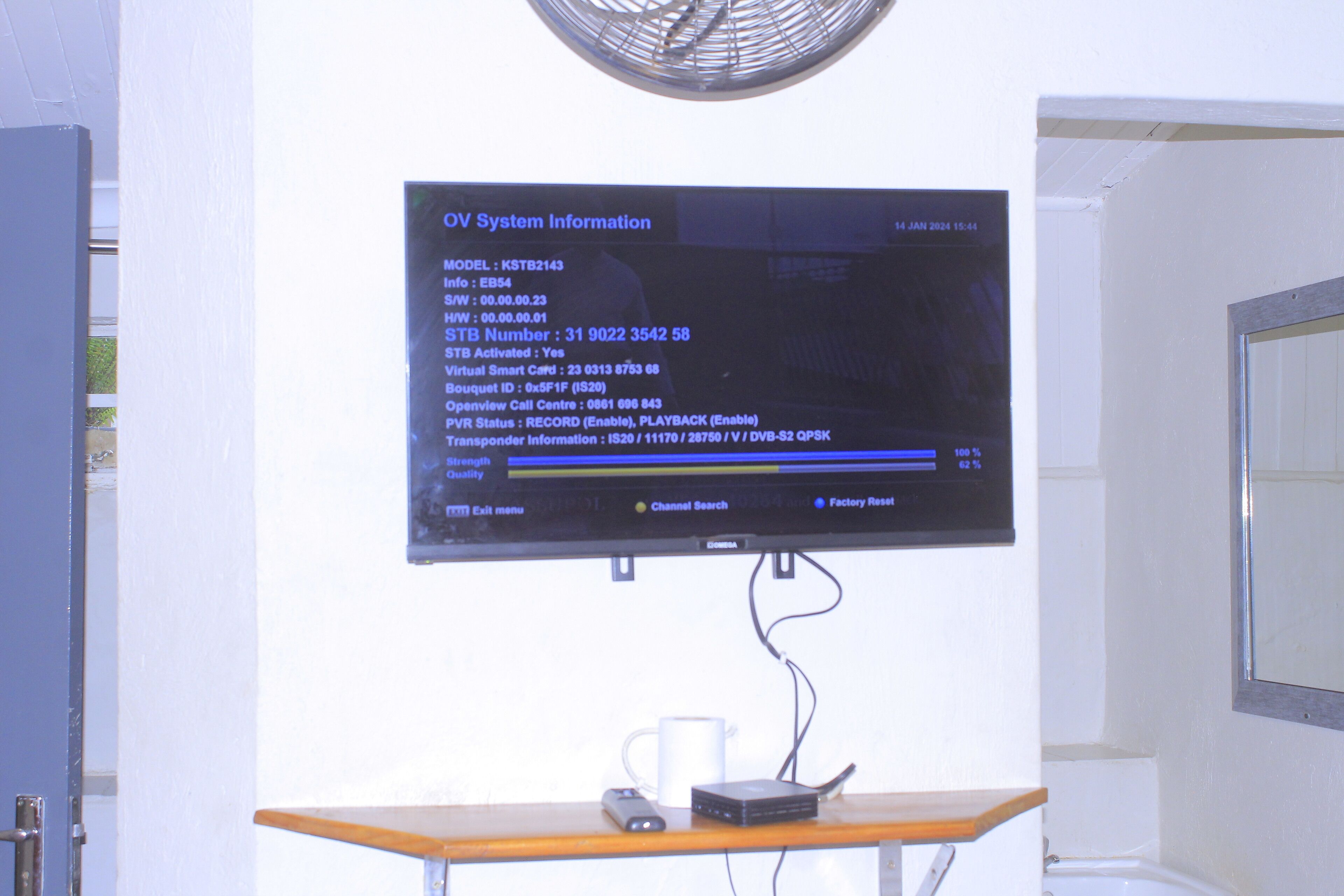 Un televisor mostrando información del sistema en una habitación con una mesa de madera, una taza blanca y un ventilador.