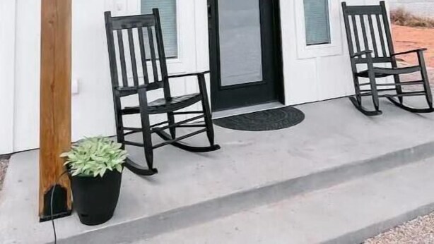 Un petit bâtiment blanc avec une porte noire, deux chaises noires et une plante en pot.