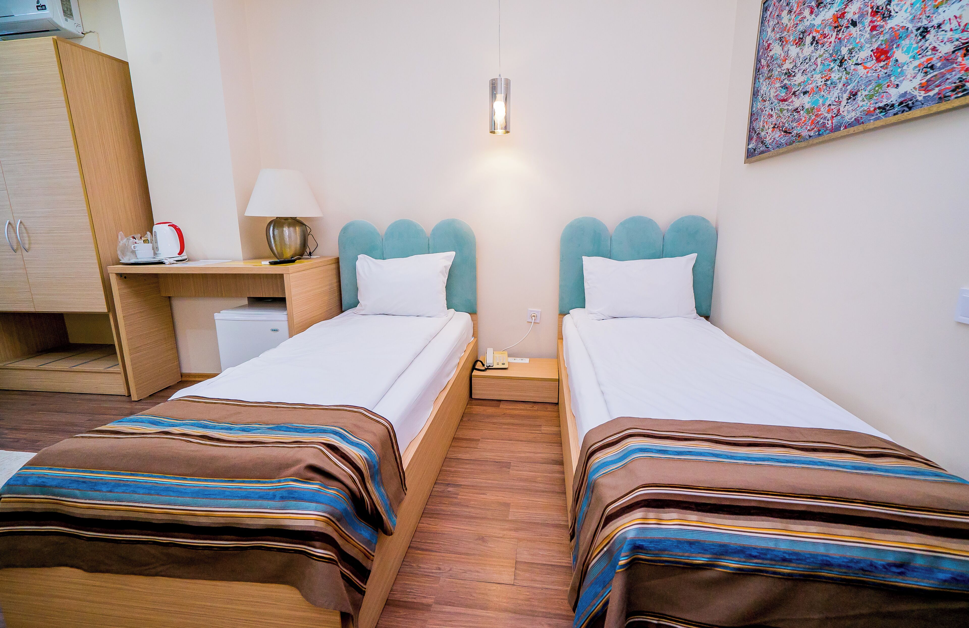A hotel room with two beds, a desk, a lamp, a painting, and a wooden wardrobe.