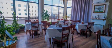 Un espace repas comprenant plusieurs tables et chaises, de grandes fenêtres donnant sur la ville et des plantes en pot.