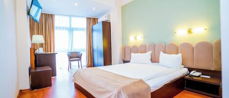 A hotel room with a large bed, a desk with a lamp, a chair, a TV mounted on the wall, and a nightstand with a phone.
