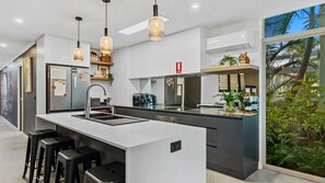 A modern kitchen with a central island, bar stools, pendant lighting, and a view of greenery outside.