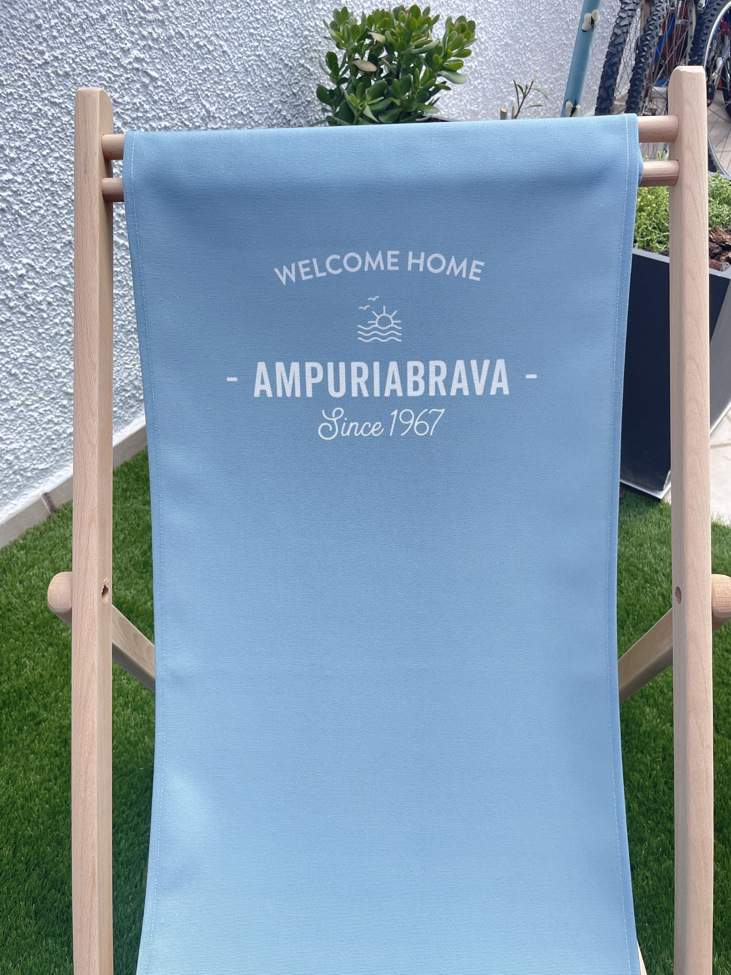 A blue towel with "WELCOME HOME AMPURIABRAVA - Since 1967" printed on it, hanging on a wooden chair.