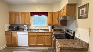 A kitchen with wooden cabinets, a dishwasher, a microwave, a stove, and a window with a view of trees. - Charming House surrounded by woods, close to Windrock and Lake Norris (Rocky Top)