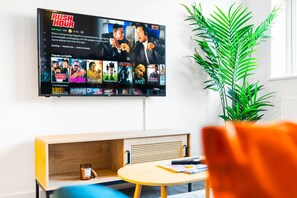 A living room with a mounted TV displaying a show, a wooden bench, a small table with a book, and a potted plant. - Luxury Apartment - Parking - WiFi - Cradley Heath
 (Cradley Heath)