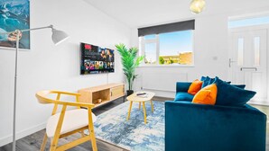 En moderne stue med en blå sofa, en træstol, et lille bord, en gulvlampe, en potteplante og et fjernsyn monteret på væggen.