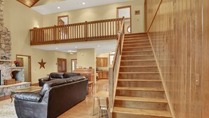 A spacious living area with a high ceiling, a wooden staircase, a leather sofa, and a stone fireplace. - Shamrock- Aunt Susie's- Raystown Lake- 7 Points (Hesston)