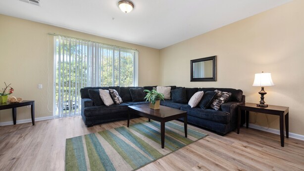 A living room with a dark-colored sofa set, a wooden coffee table, a striped rug, a lamp, and a sliding glass door leading to a balcony.