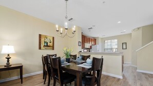 A dining area with a wooden table, chairs, and a chandelier. There is a kitchen in the background with wooden cabinets and a microwave.