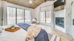 Ein geräumiges Schlafzimmer mit einem großen Bett, einem Kamin und Blick auf den Wald durch große Fenster.