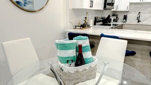 Eine moderne Küche mit weißen Schränken, einer weißen Arbeitsplatte und einem Glastisch mit zwei weißen Stühlen. Auf dem Tisch steht ein Korbwicker-Korb mit einer Flasche und Handtüchern.