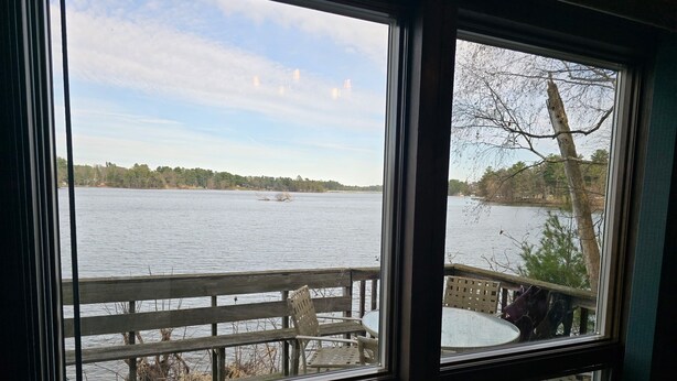 Vue depuis une chambre dotée d’une grande fenêtre donnant sur un lac, une terrasse en bois et un espace salon extérieur.