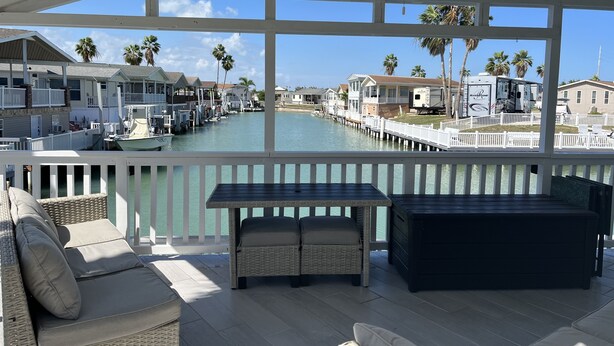 A waterfront balcony with wicker furniture, a ceiling fan, and a view of a marina with boats and houses.