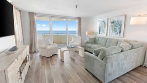 Una sala moderna con un sofá grande de color gris, un sillón blanco y una mesa de centro. Se ve una ventana grande con vistas al mar.