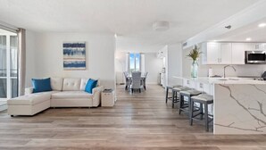 Una sala de estar moderna con un sofá modular, una zona de comedor y una cocina con armarios blancos y encimera de mármol.