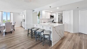 Una cocina moderna con armarios blancos, una isla de mármol y un comedor con sillas de mimbre.