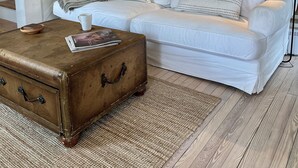 Una sala de estar con un sofá blanco, un baúl marrón, una escalera y un estante de pared con adornos.