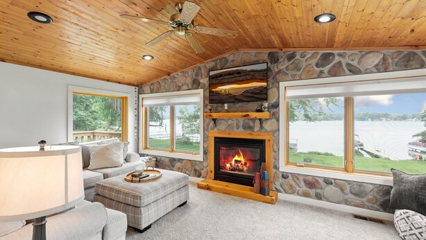 A cozy living room with a stone fireplace, large windows overlooking a lake, and a ceiling fan. - Lakeside Retreat- 7beds/4bd/2ba! Family and pet friendly!  (Zimmerman)