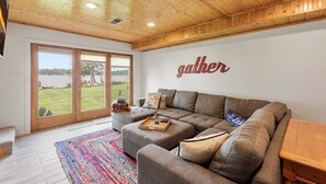 A modern living room with a large sectional sofa, a wooden ceiling, and a view of a green landscape through glass doors. - Lakeside Retreat- 7beds/4bd/2ba! Family and pet friendly!  (Zimmerman)