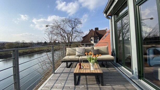 Eine Holzterrasse mit einer Couch und einem Tisch, mit Blick auf einen Fluss und ein Gebäude mit rotem Dach.