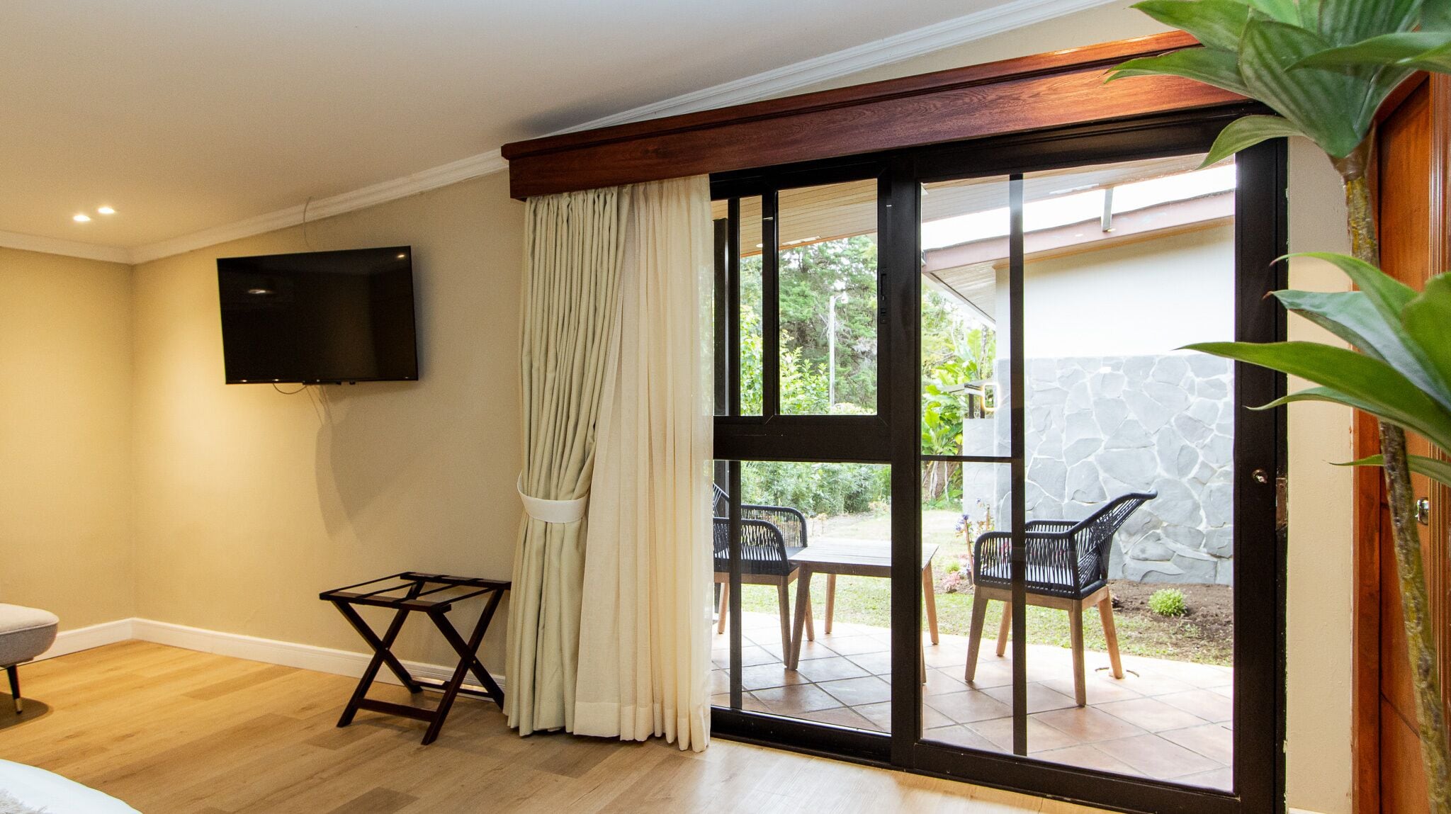 A room with a TV mounted on the wall, a sliding glass door leading to an outdoor patio with chairs and a table, and a small wooden stool.