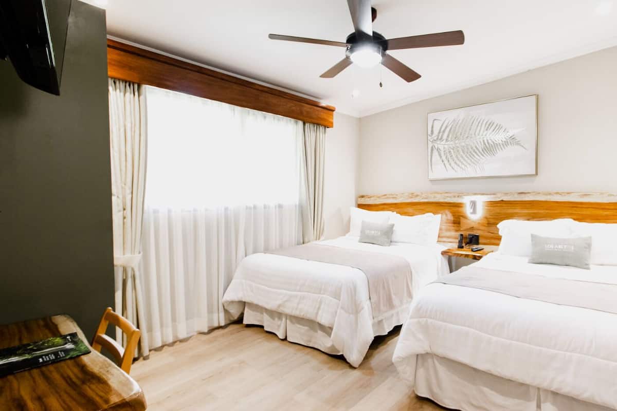 Une chambre d’hôtel avec deux lits, une tête de lit en bois, un ventilateur de plafond, une table de chevet avec une lampe et une œuvre d’art représentant une feuille, encadrée, accrochée au mur.
