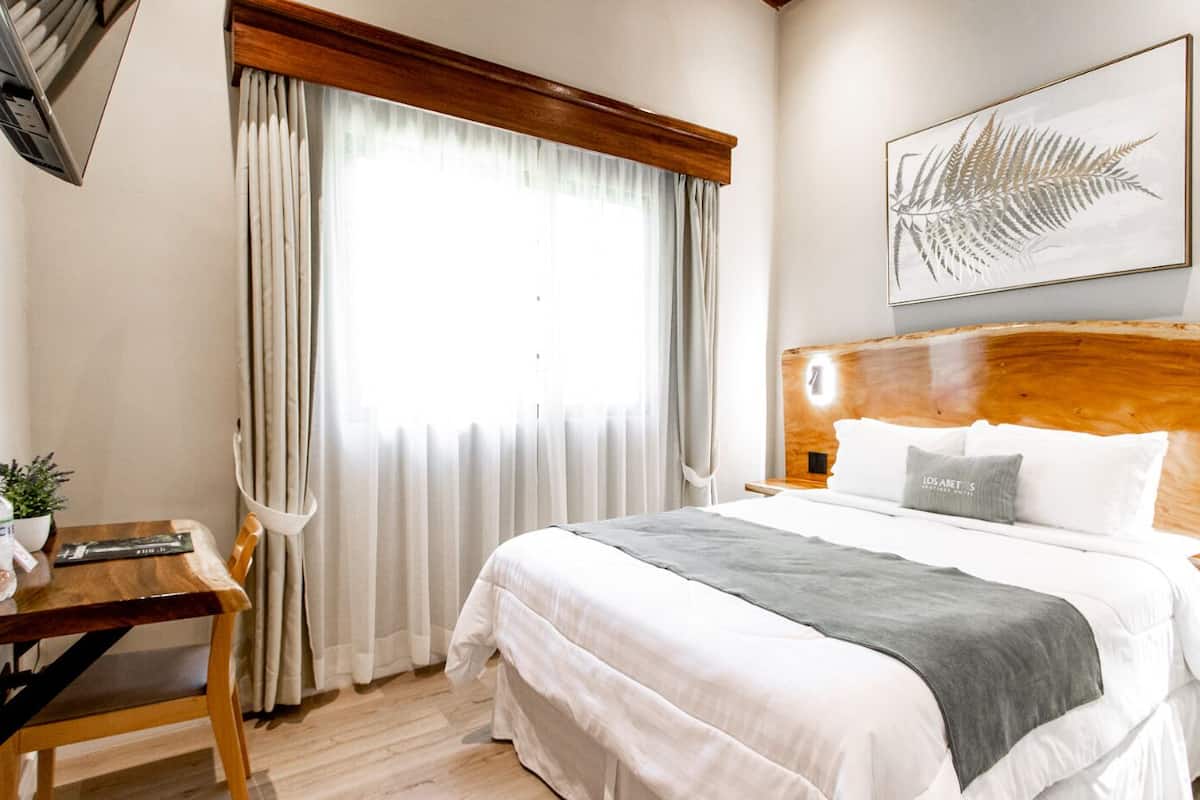 Une chambre d’hôtel avec un grand lit, une tête de lit en bois, une table de chevet avec une lampe, un bureau avec une chaise et une fenêtre avec des rideaux.
