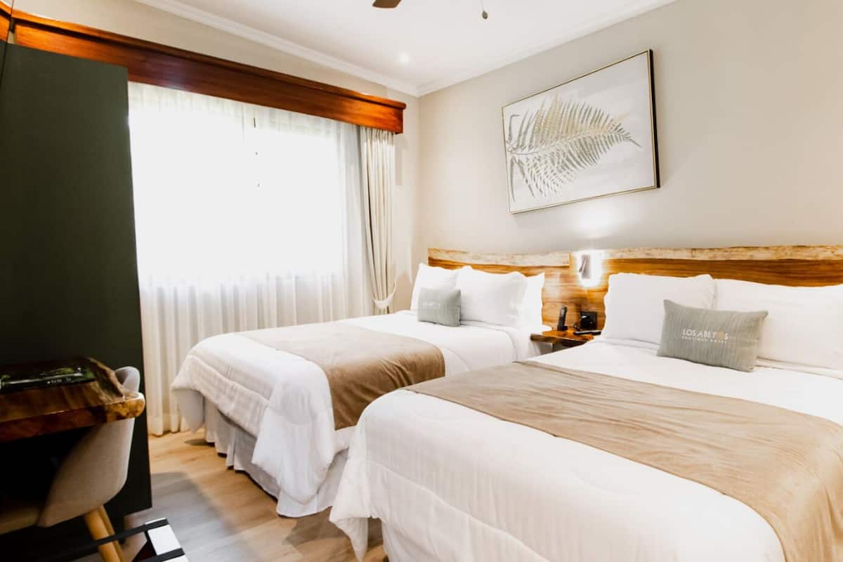Une chambre d’hôtel avec deux lits, une tête de lit en bois, un bureau et une grande fenêtre avec des rideaux.