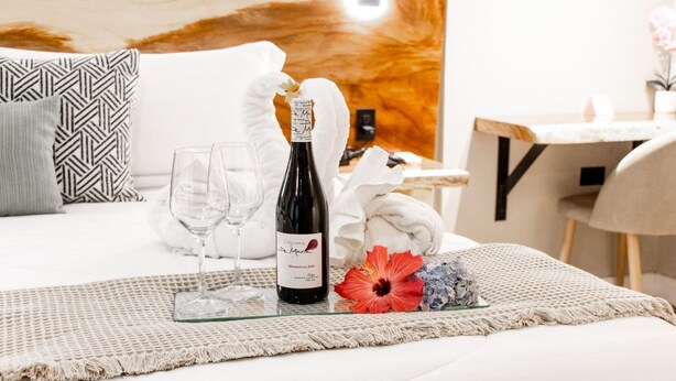 Una cama bien hecha con una almohada blanca, una bandeja de vidrio con una botella de vino, dos copas y una flor de hibisco roja.