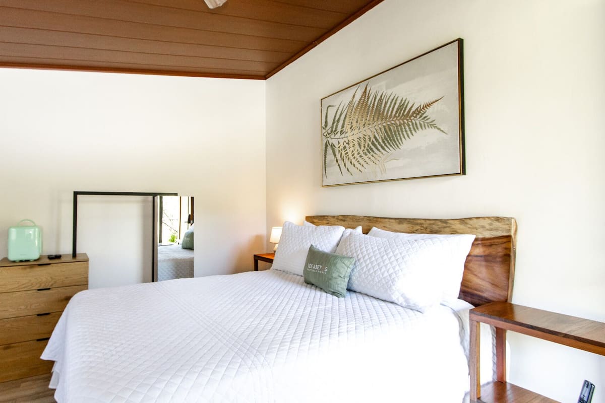 Une chambre avec un plafond en bois, un lit recouvert de linge de lit blanc, une table de chevet avec une lampe, une commode en bois et une œuvre d’art représentant une fougère, encadrée, accrochée au mur.