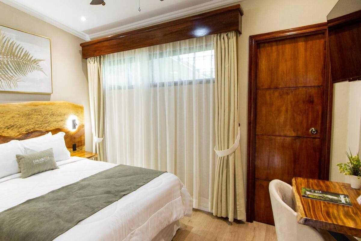 Une chambre d’hôtel comprenant un lit, un bureau, une chaise, une tête de lit en bois, une grande fenêtre avec des rideaux et une plante fixée au mur.