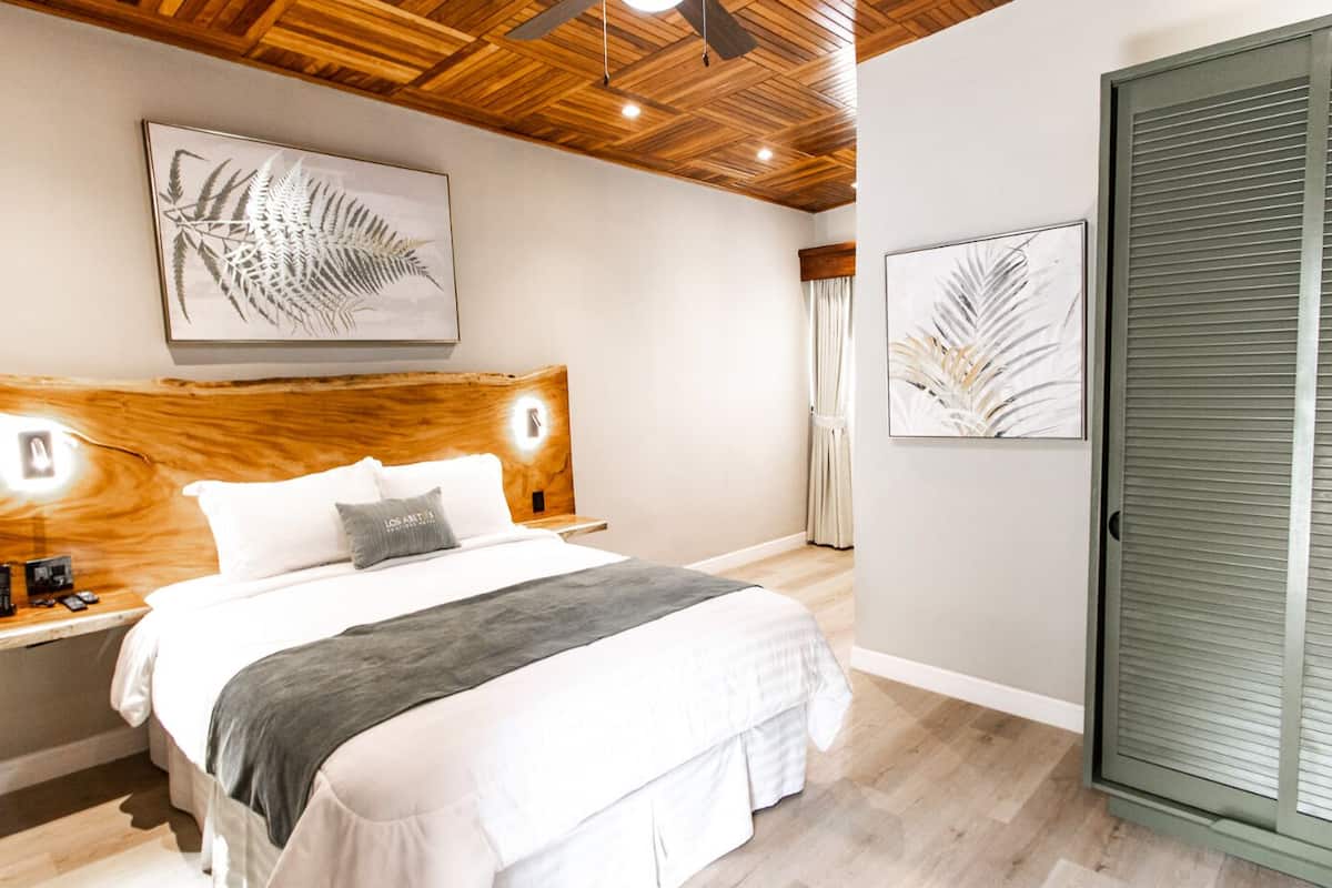 Une chambre à coucher avec une tête de lit en bois, un lit recouvert d’une literie blanche, un plaid gris et deux œuvres d’art représentant des feuilles, encadrées et accrochées aux murs.