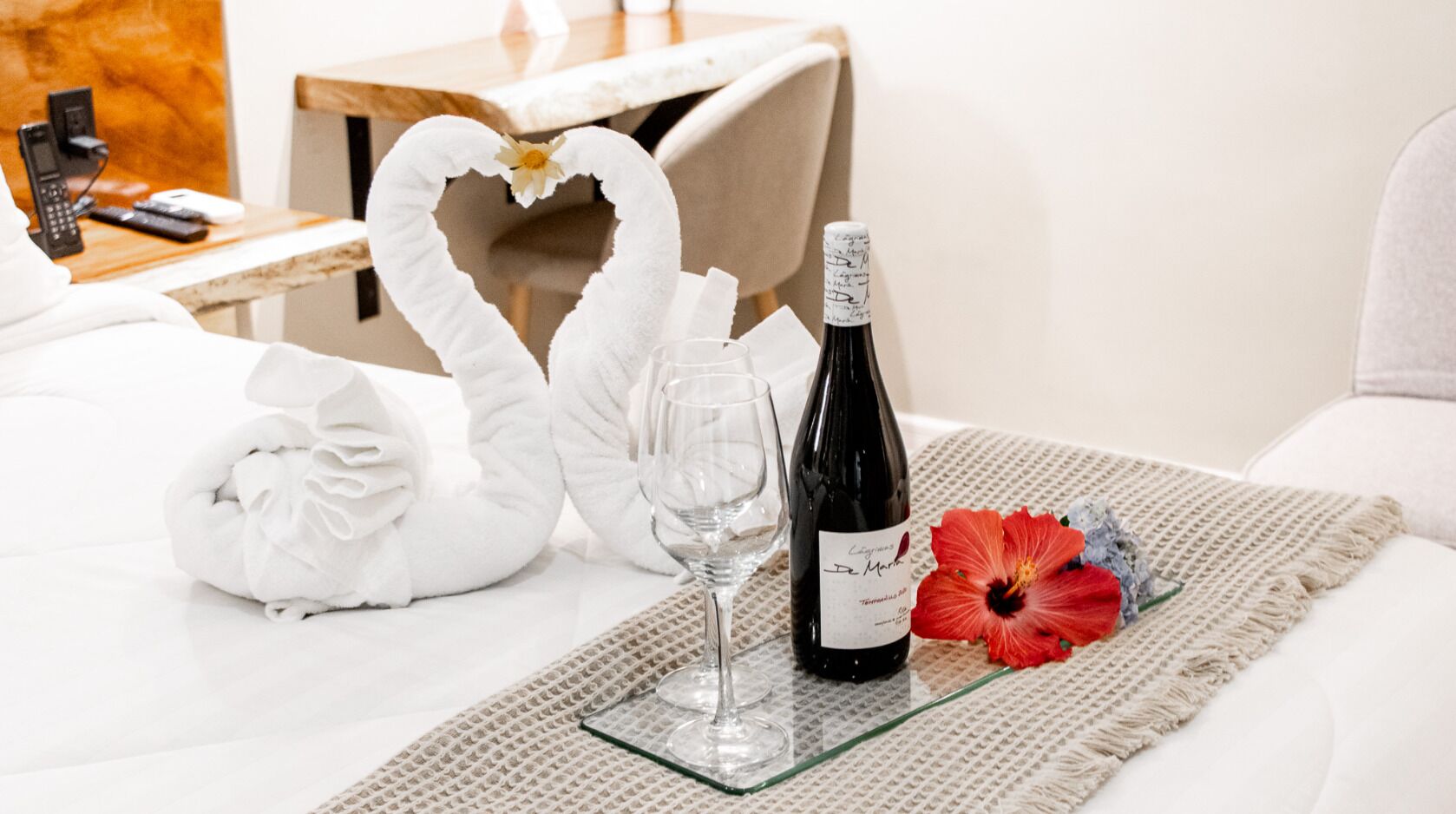 A neatly made bed with a glass tray holding a bottle of wine, two glasses, and a decorative swan towel.