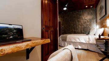 A bedroom with a bed, a wooden nightstand, a laptop on a wooden table, and a stone wall.