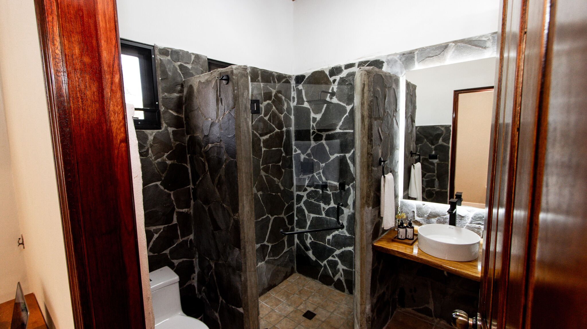 Een badkamer met een douchecabine van natuursteen, een toilet en een wastafel met spiegel.