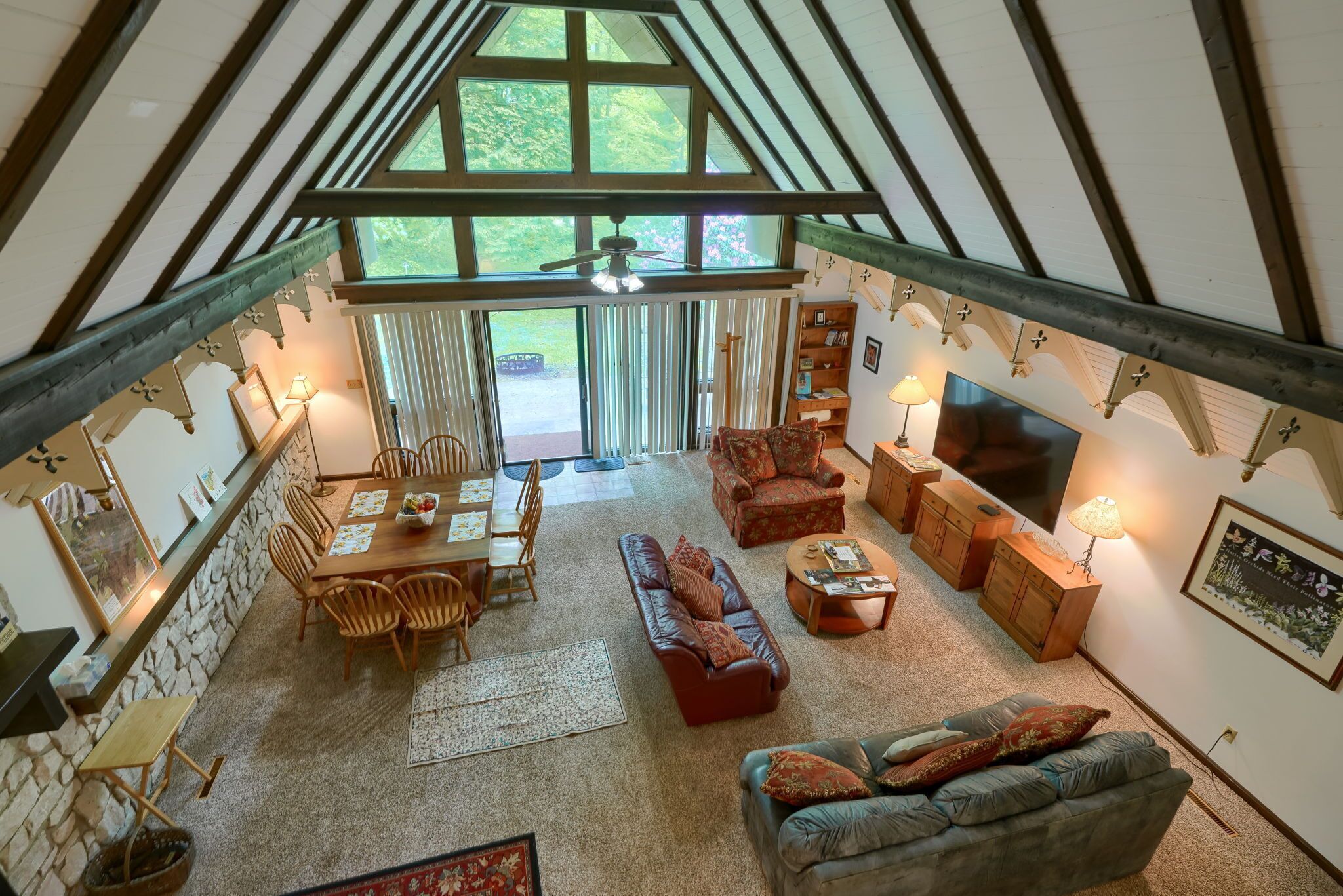 A spacious interior with a high ceiling, a dining area, a living room with a sofa and TV, and a view of the outdoors through large windows.