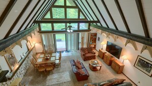 A spacious interior with a high ceiling, a dining area, a living room with a sofa and TV, and a view of the outdoors through large windows. - Mercer House in Hocking Hills (Sugar Grove)