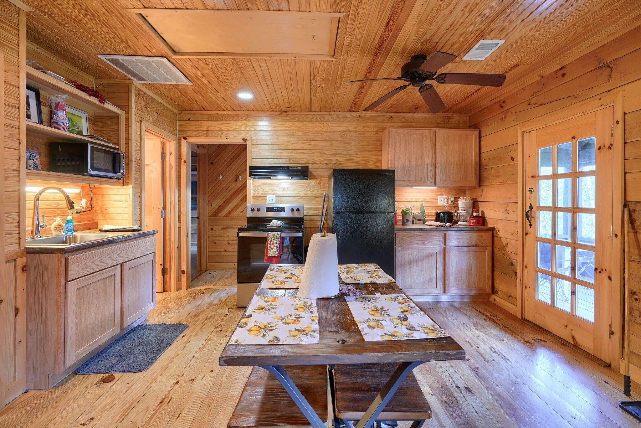 Interior de una cabaña de madera con cocina, muebles de madera, un ventilador de techo y una mesa de comedor con individuales.