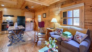 L’intérieur chaleureux d’un chalet avec un plancher en bois, une table à manger, un fauteuil à bascule, un canapé et un coin cuisine équipé d’un réfrigérateur et d’une cuisinière.