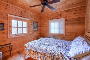 Une chambre dans une cabane en bois, avec un lit, un ventilateur de plafond et deux fenêtres.