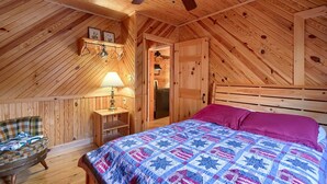 Une chambre mansardée chaleureuse avec un plafond en pente en bois, un lit recouvert d’une couverture colorée, une chaise et une petite table avec une lampe.