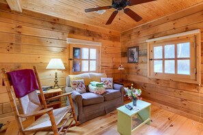 Ein gemütliches Blockhaus-Interieur mit Holzbalkendecke, einem Schaukelstuhl, einer Couch, einem kleinen Tisch mit Lampe und zwei Fenstern.