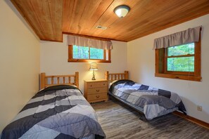 A bedroom with two beds, wooden headboards, a nightstand, a lamp, and two windows with curtains.