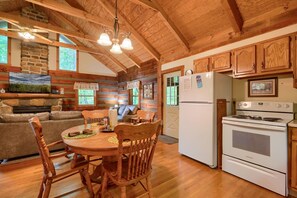 A cozy cabin interior with a dining area, wooden furniture, a stone fireplace, and a kitchen with a stove and refrigerator.