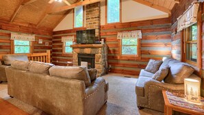 A log cabin interior with a stone fireplace, wooden walls, and a ceiling fan. - Chickasaw in Hocking Hills (Logan)