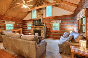 A log cabin interior with a stone fireplace, wooden walls, and a ceiling fan.