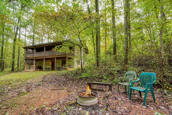 A cabin with a deck, a fire pit, and two chairs in a forested area. - Chickasaw in Hocking Hills (Logan)
